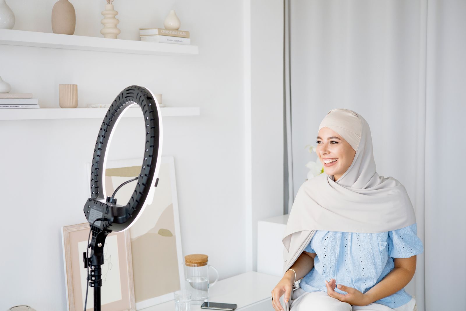 Smiling creator in a hijab recording content with a ring light and phone