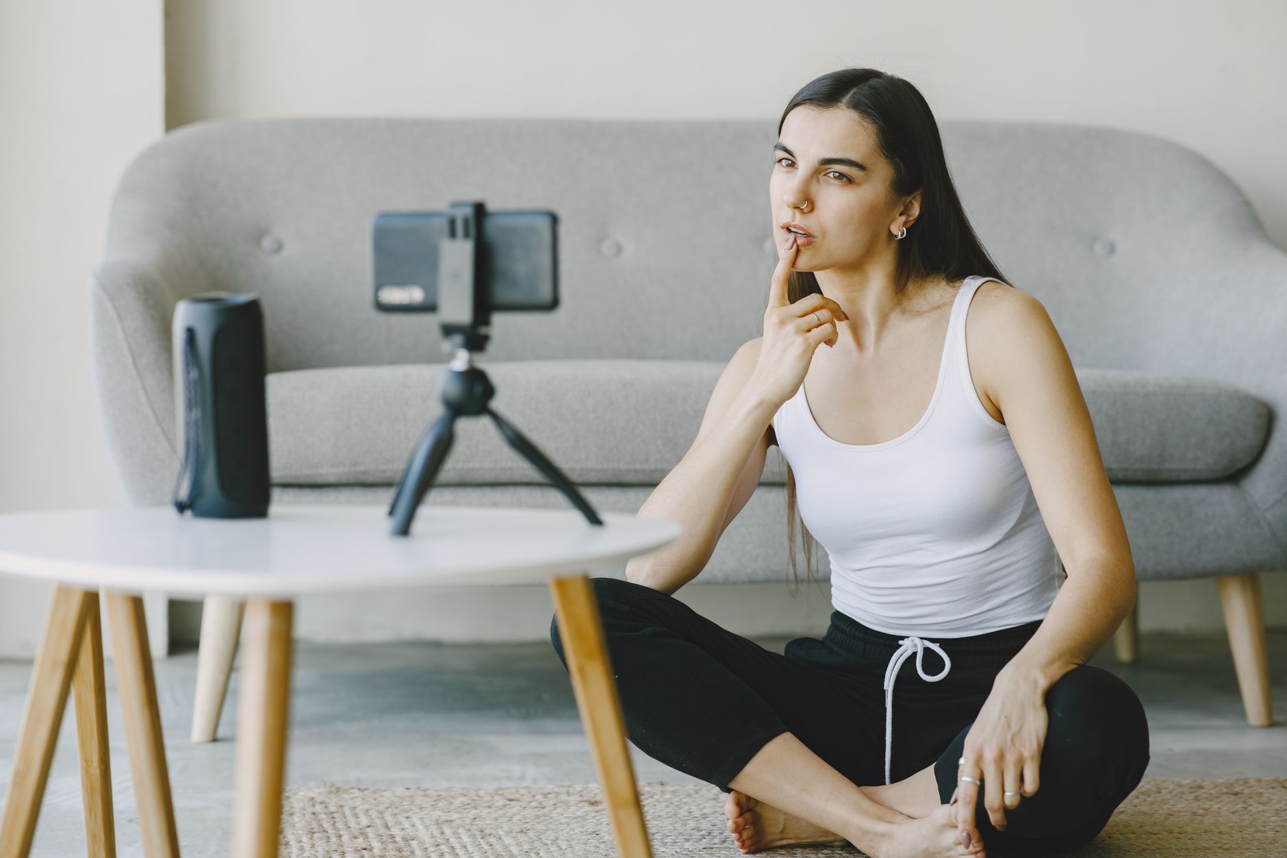 A woman sits cross legged in a living room recording video on a smartphone mounted on a small tripod, with a portable speaker nearby.