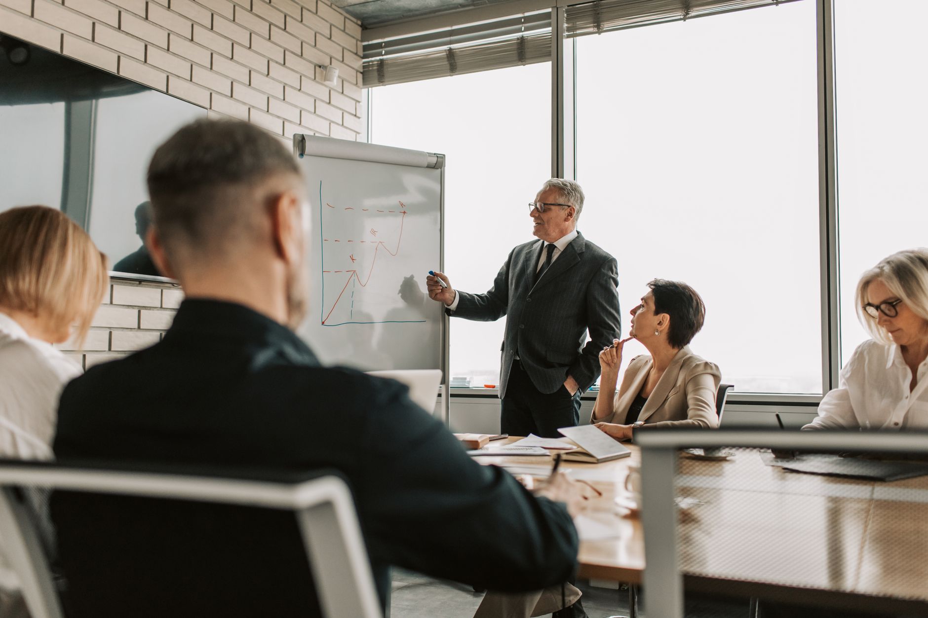 A senior executive presents a hand drawn growth chart to colleagues in a meeting room while one listener looks skeptical.