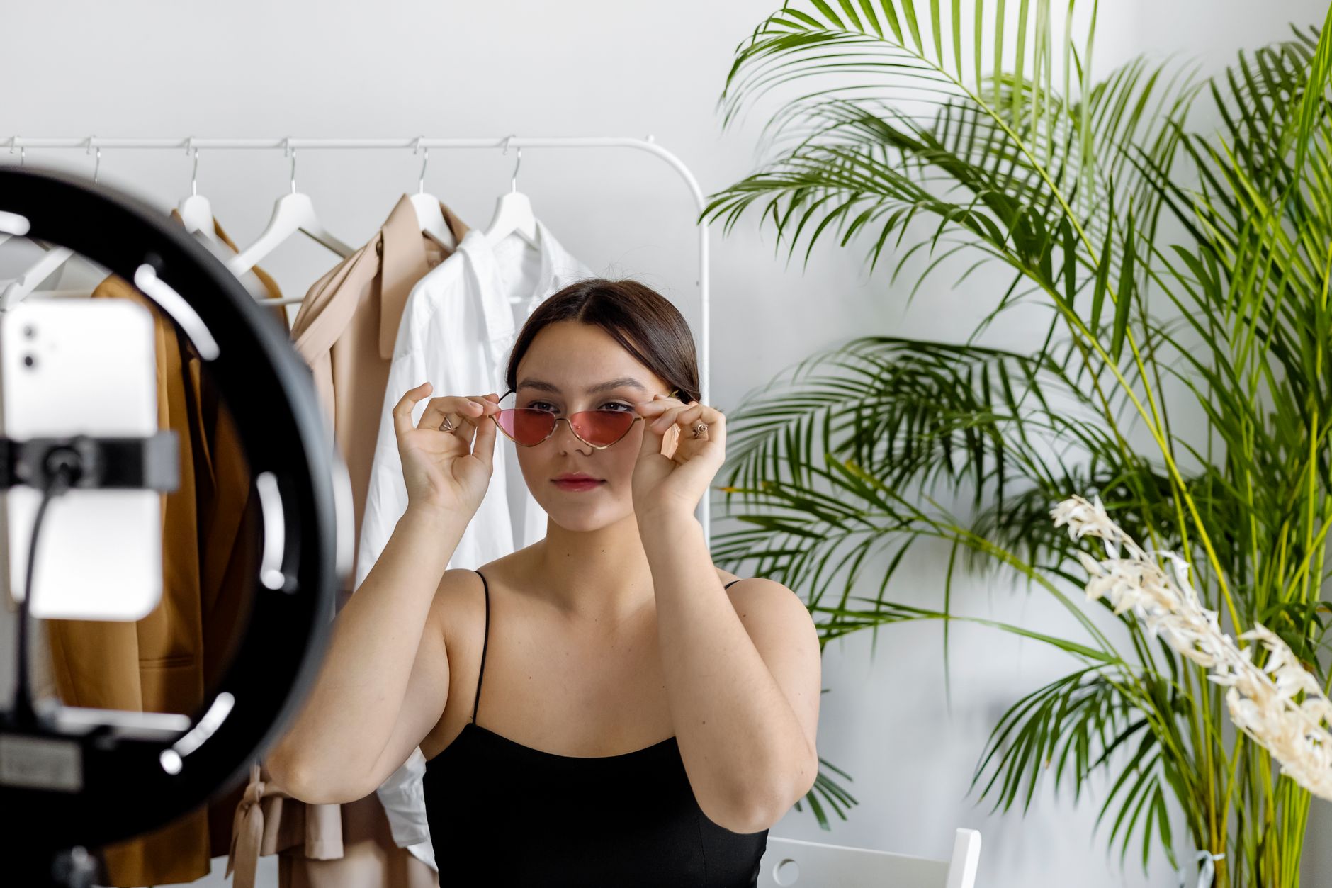 Content creator trying on sunglasses while filming in front of a ring light and smartphone, with a clothing rack in the background.