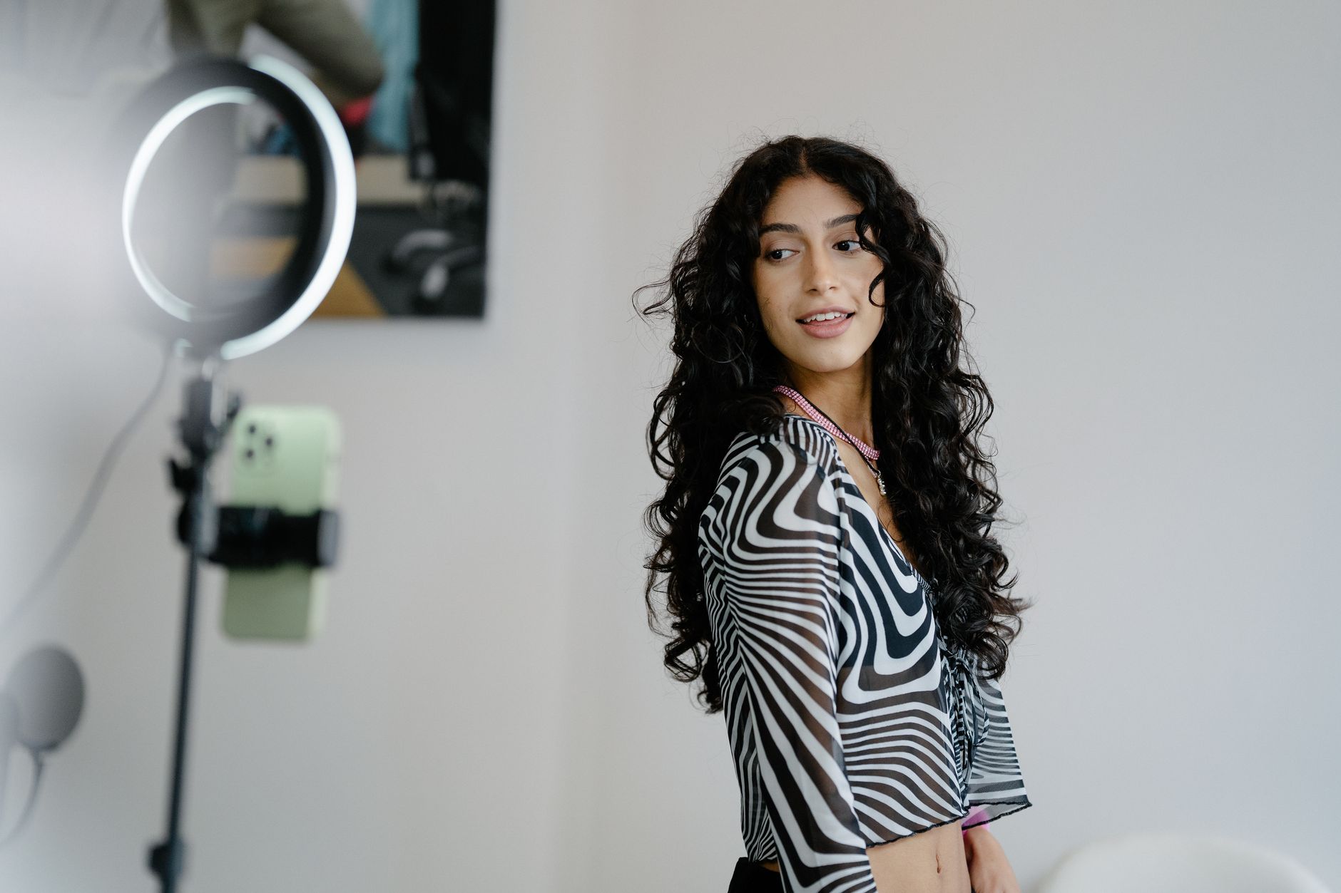 Woman with curly hair filming herself using a smartphone on a ring light stand in a plain room