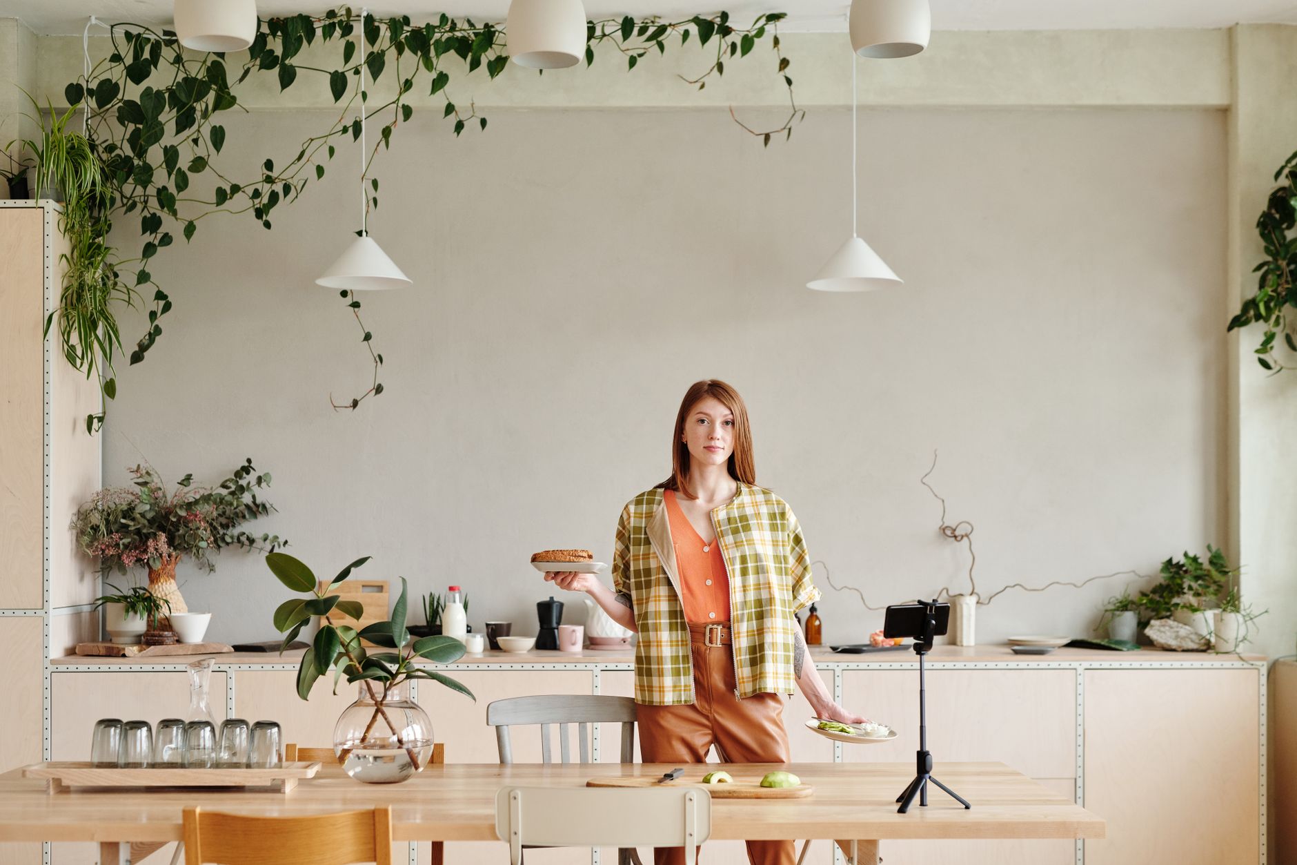 A woman films cooking content for Instagram using a phone on a tripod in her kitchen, holding plates to show on camera.