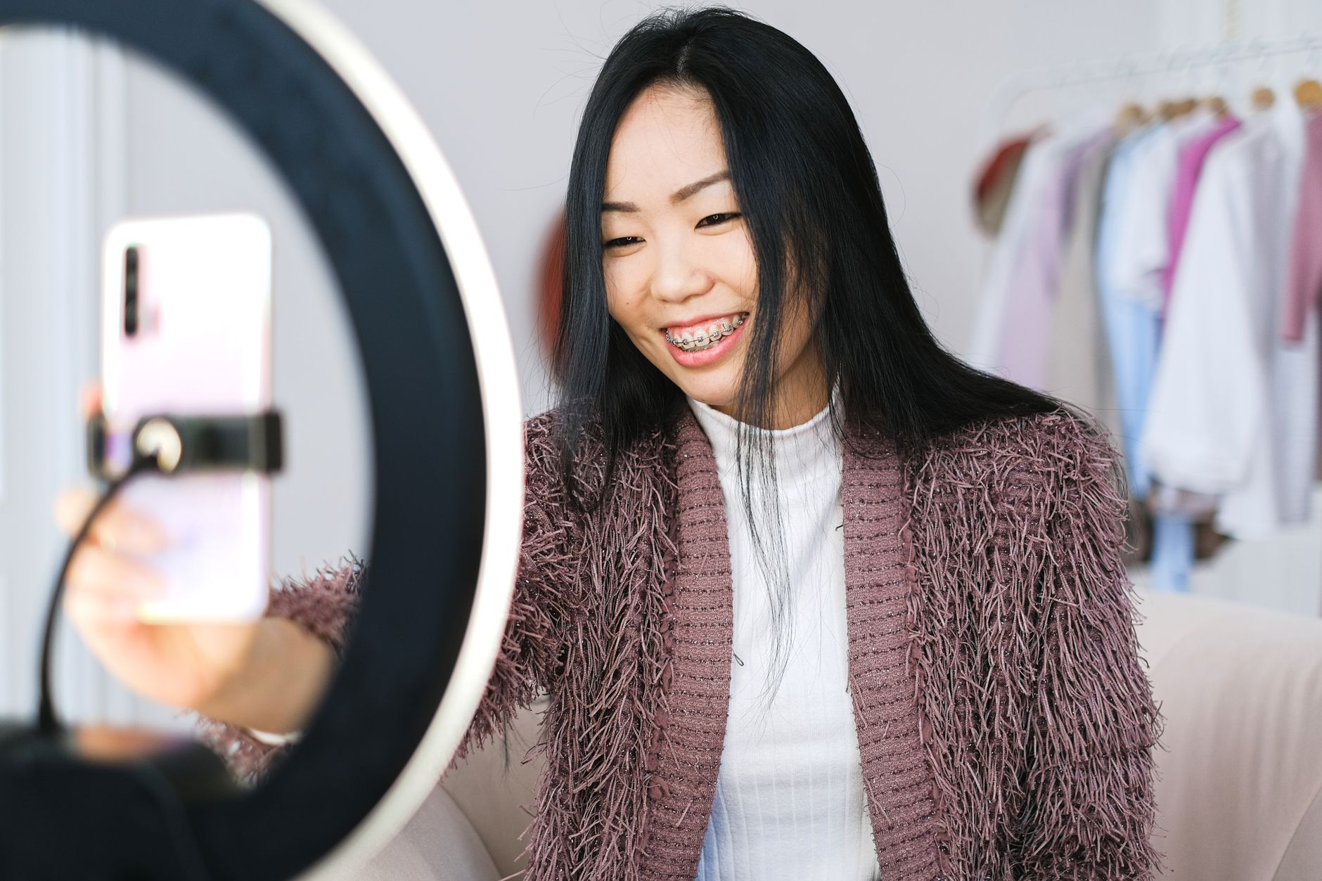 Smiling content creator holding a smartphone beside a ring light with a clothing rack in the background