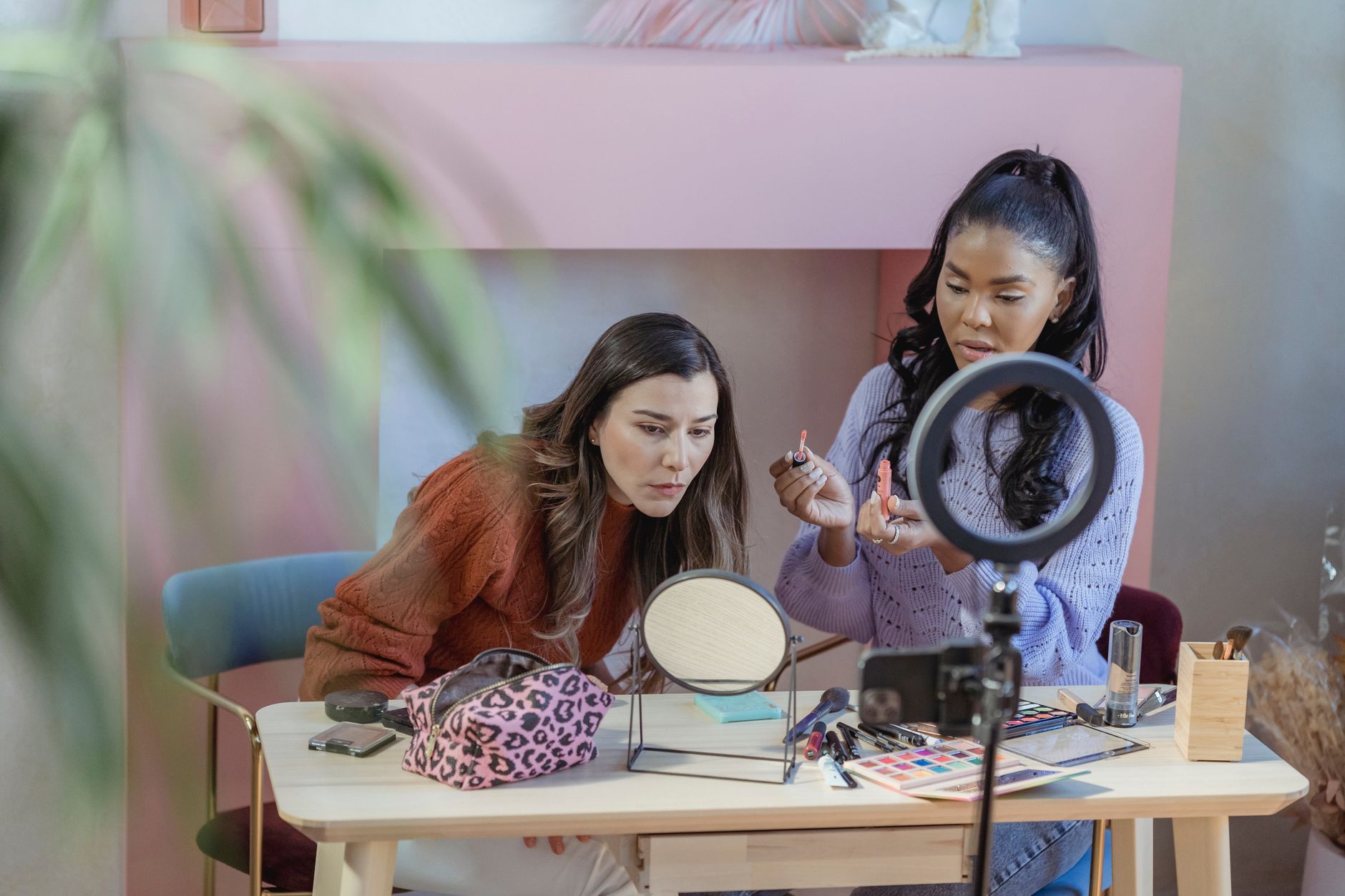 Two women recording a beauty tutorial together at a desk with a ring light, mirror, and cosmetics