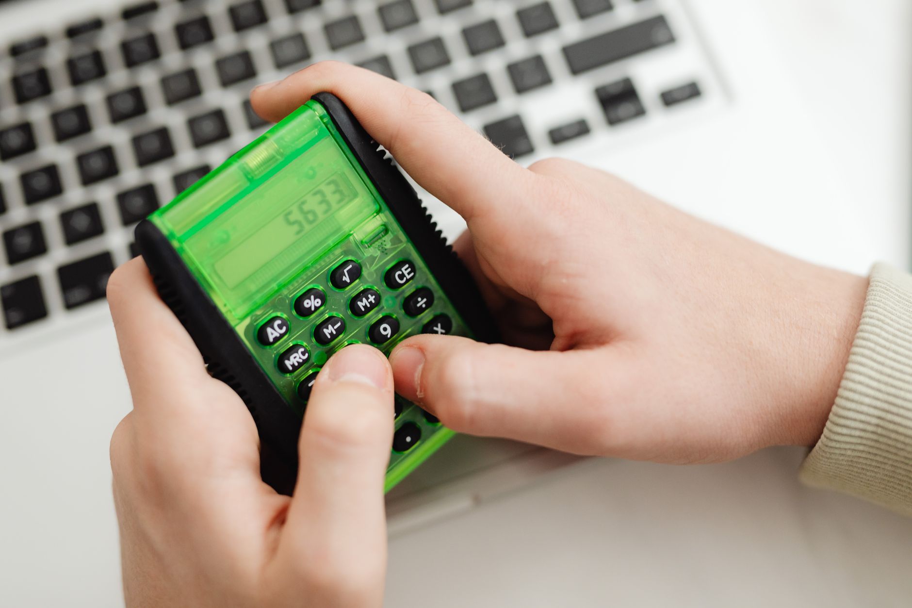 A hand uses a green calculator next to a laptop keyboard.