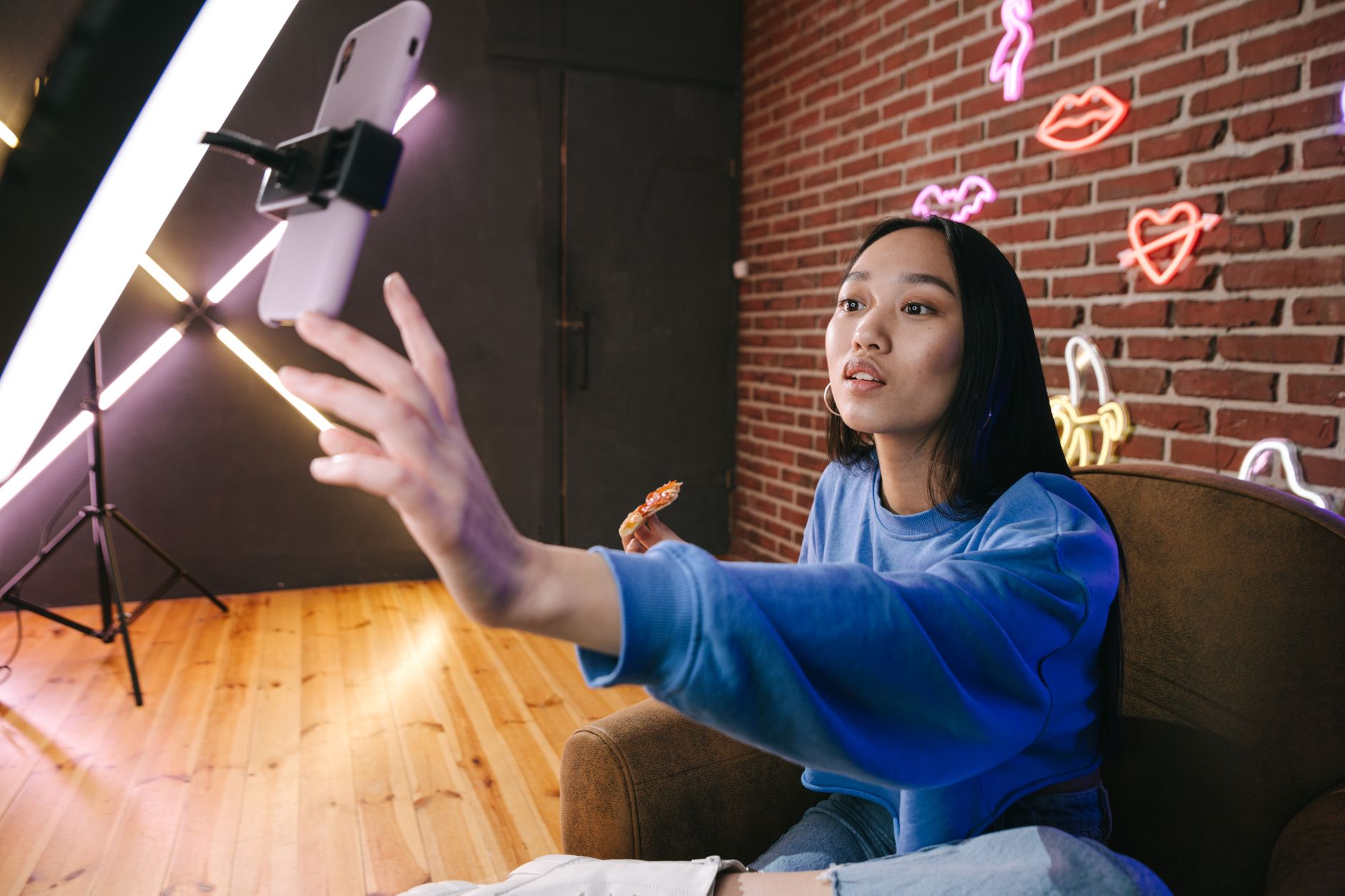 A creator films herself on a smartphone using a ring light, with neon sign decorations on a brick wall behind her